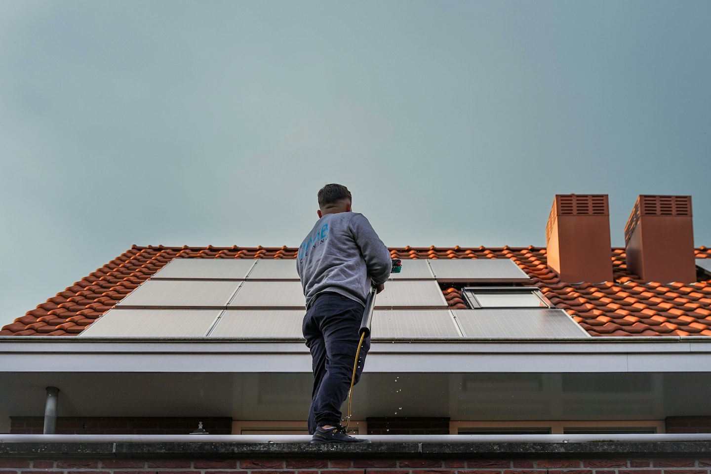 Worker cleans solar panels on an orange tiled roof with a long brush.